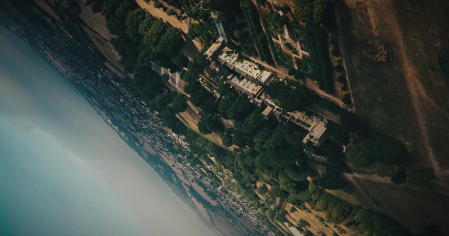 A cinematic aerial shot slowly tilts and rotates over a grand historic castle estate nestled in the lush, rolling English countryside