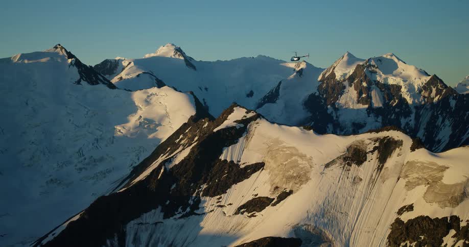 A helicopter majestically soars over a vast, snow-covered mountain range bathed in the warm light of sunrise
