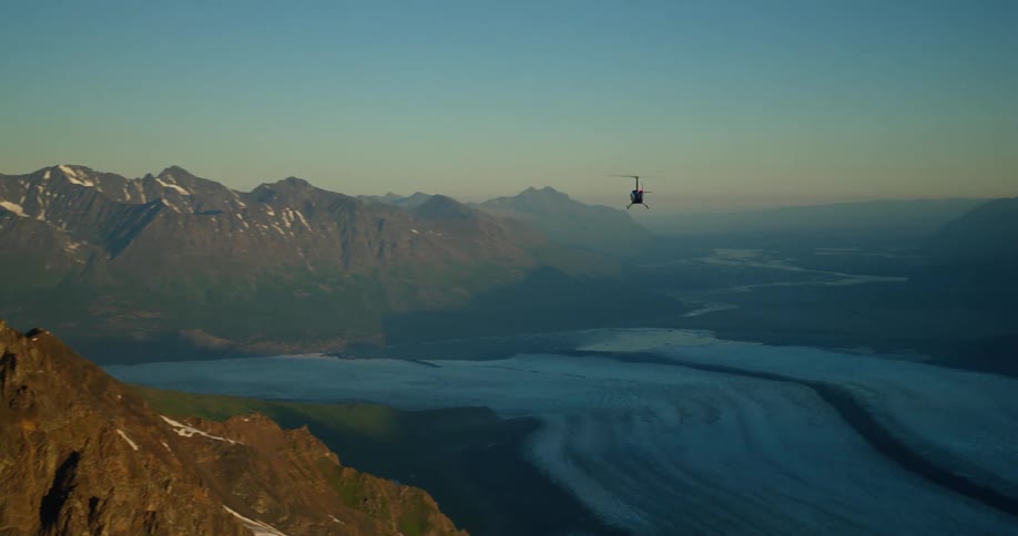 A helicopter flies past a majestic, rocky mountain peak revealing a vast glacial valley below at sunset