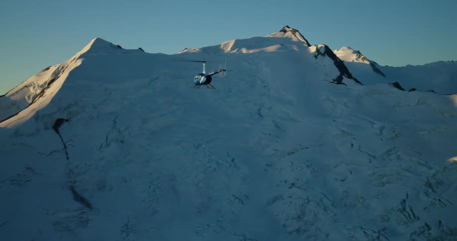A helicopter soars majestically over a vast, snow-covered glacial mountain range illuminated by the golden light of sunset