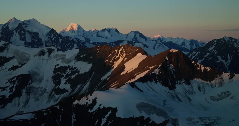 A serene, static shot captures the warm glow of sunrise illuminating the peaks of a vast, snow-covered mountain range