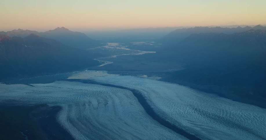 A serene, high-altitude aerial view of a massive glacier carving its way through a vast mountain valley at dusk