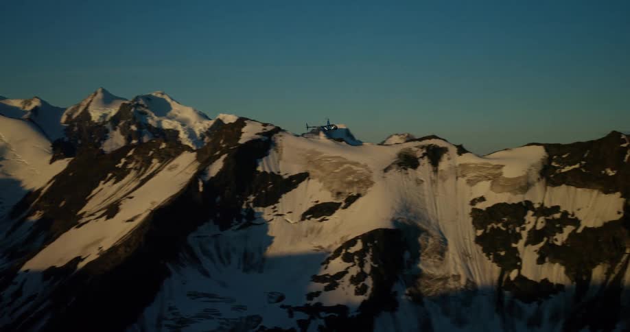 A helicopter soars over majestic, snow-capped mountains and glaciers under a clear sky at sunset