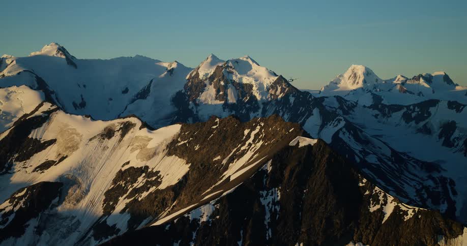 An aerial shot follows a small plane as it soars over a majestic snow-capped mountain range at sunrise