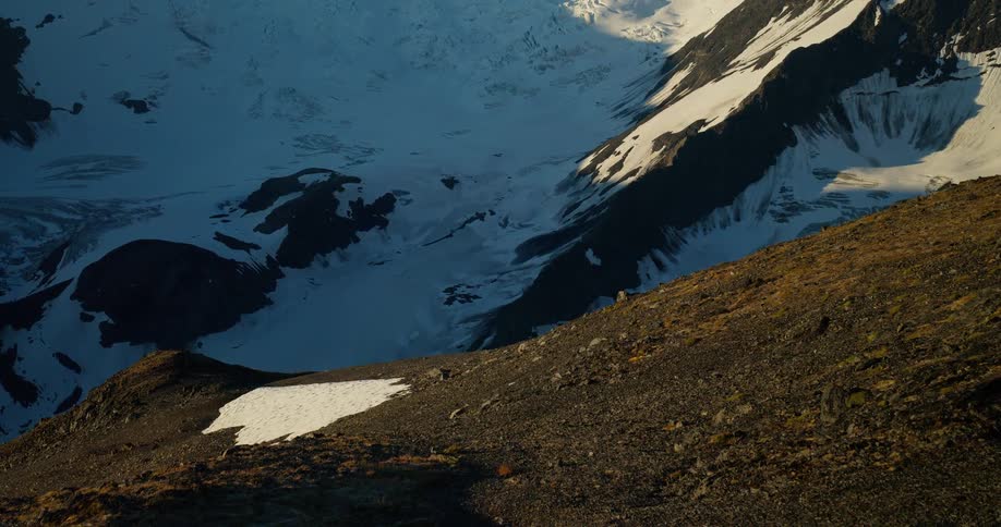 A time-lapse captures the morning sun casting long shadows across a rugged mountain landscape and a massive glacier below