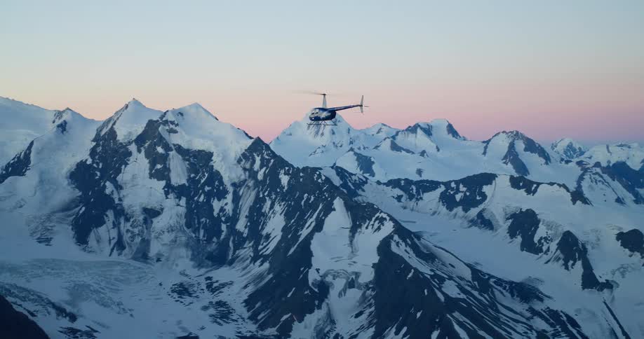 A helicopter flies gracefully over a stunning, snow-covered mountain range during a beautiful sunset