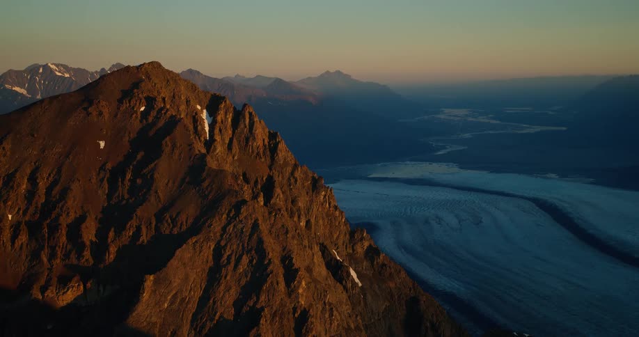 A dramatic mountain peak catches the golden sunrise light, overlooking a massive, winding glacier in the vast valley below