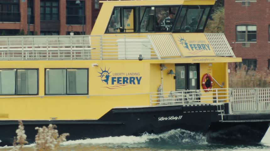 A yellow and black Liberty Landing Ferry boat travels down a river in slow motion with brick buildings along the shoreline