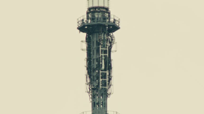 A slow, upward tilt reveals the intricate metal lattice and spire of a tall communications tower against a plain sky