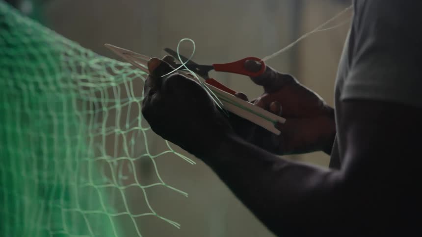 Close up of a silhouetted fisherman skillfully mending a large green fishing net by hand in a dimly lit room