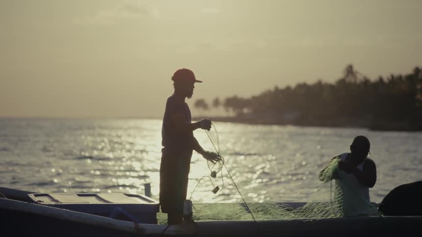 Two fishermen are silhouetted against the golden hour sun as they work together preparing their fishing nets on a boat