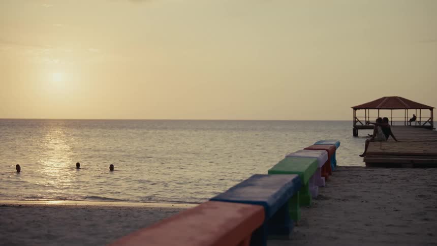 A peaceful beach scene unfolds at sunset as people swim in the calm ocean and a couple sits on a pier
