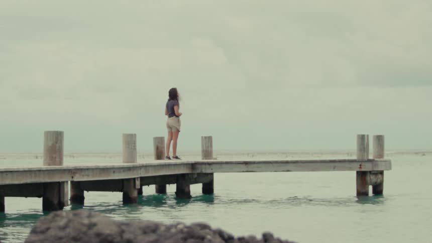 A woman stands alone on a tranquil wooden pier, gazing out at the calm ocean on a cloudy day