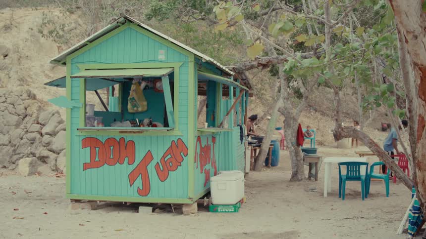 A colorful 'Dom Juan' food shack sits on a sandy tropical beach as people go about their day in the background