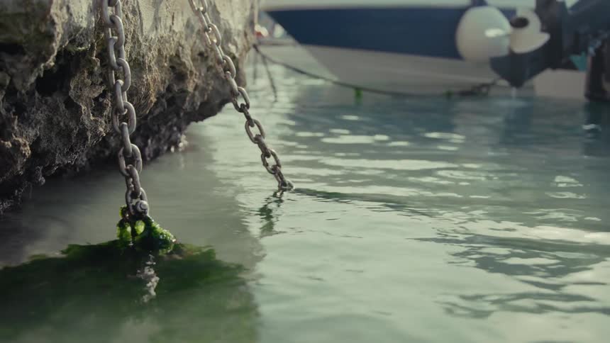 Mossy chains hanging from a stone pier gently move in the calm harbor water as a boat floats in the background