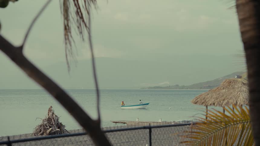 A tranquil view of a small fishing boat floating on calm tropical waters, with a slow focus pull to the foreground palm leaves