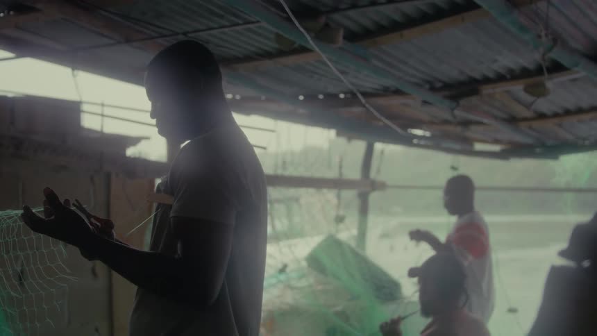 Silhouetted fishermen meticulously repair their green fishing nets in a rustic waterside shed