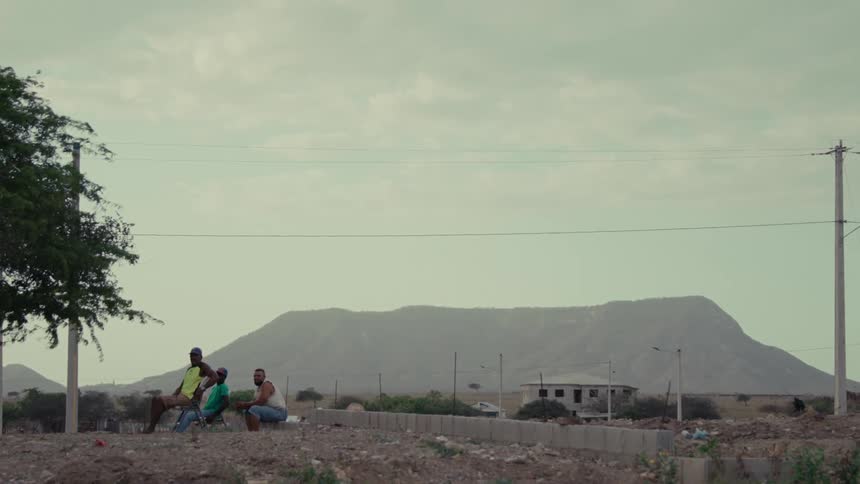 Three men gather and chat in a quiet, rural landscape with a hazy mountain in the background