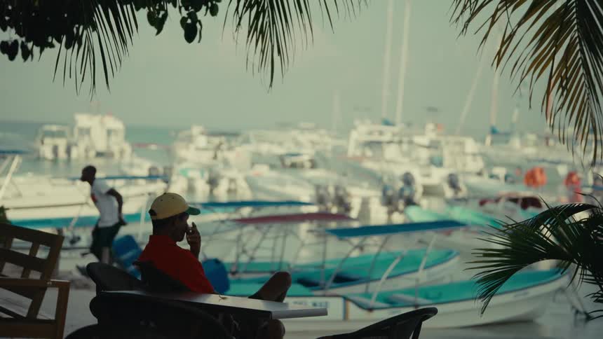 A man in a red shirt talks on the phone while looking out at a tropical marina full of boats