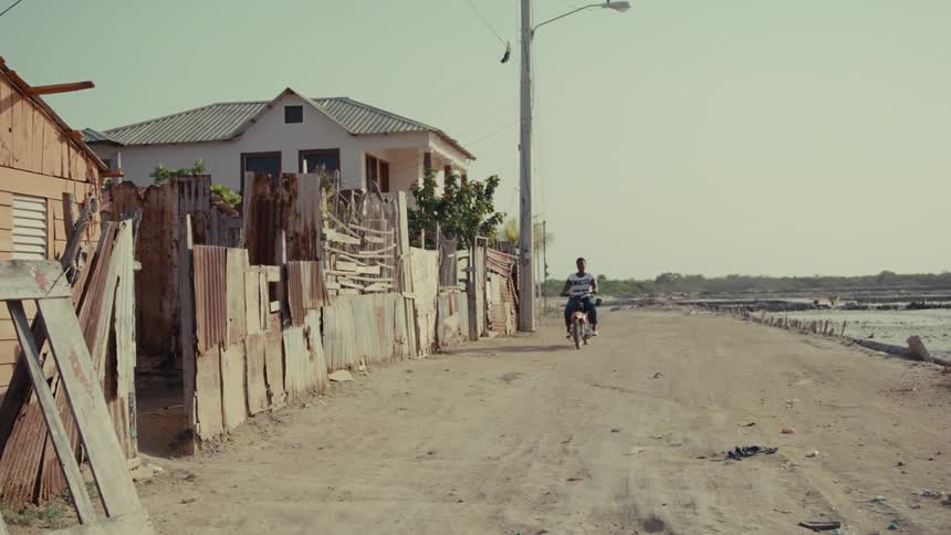 A man rides a motorcycle down a dusty dirt road in a rustic village during the day
