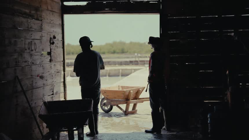 Two silhouetted workers stand in the doorway of a rustic wooden shed, conversing while looking out at a bright salt flat