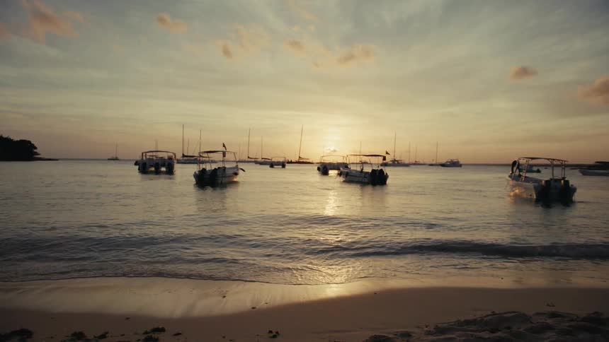 A tranquil ocean bay with anchored boats basks in the golden light of a setting sun as viewed from the sandy shoreline
