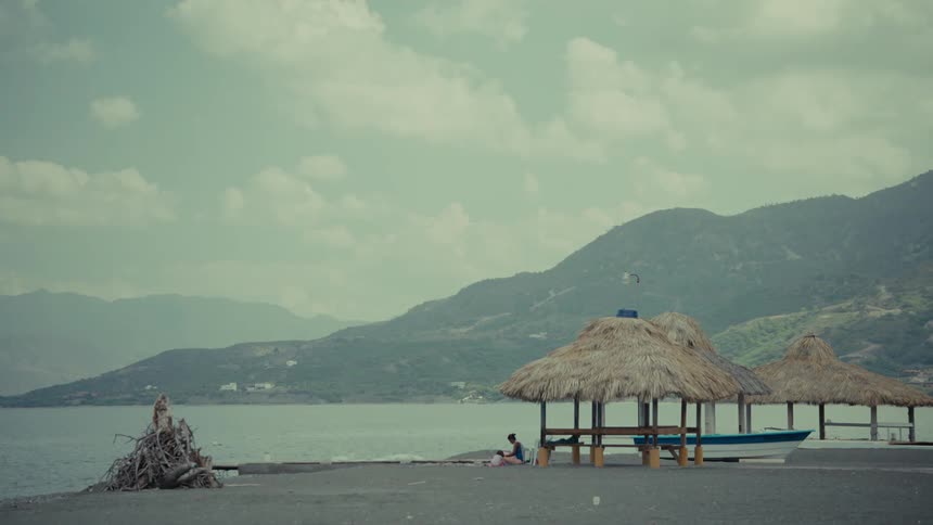 A serene, wide shot of a person sitting under a thatched-roof hut on a quiet shoreline with mountains in the background