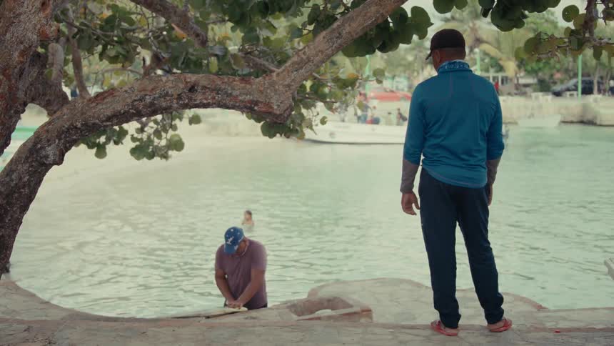 A man stands on a stone ledge watching another man clean a freshly caught fish in the shallow waters of a tropical bay