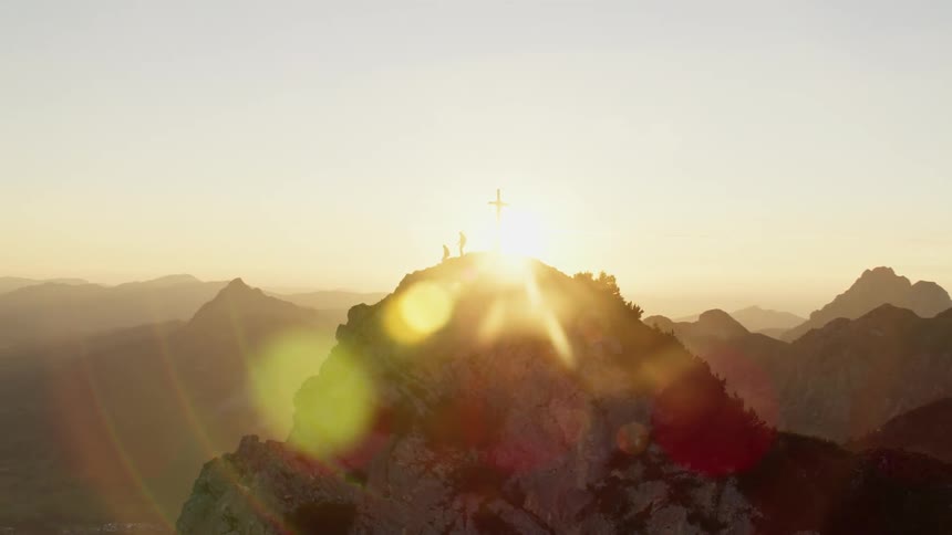 Two people standing by a cross at the top of a rocky mountain peak