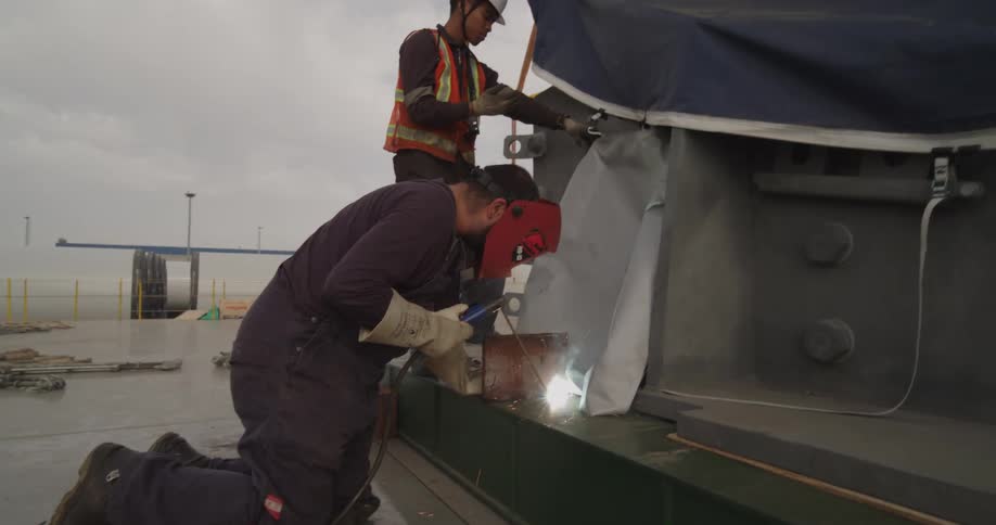 A skilled worker wearing a protective mask and gloves kneels as he meticulously welds a large metal structure at an industrial site