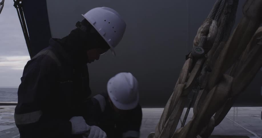Two seamen in hard hats and overalls work together to secure heavy rigging on the deck of a ship at sea