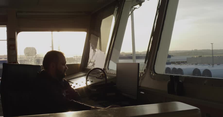 A ship's captain works at a computer console on the bridge of a vessel while docked in port during a golden sunset