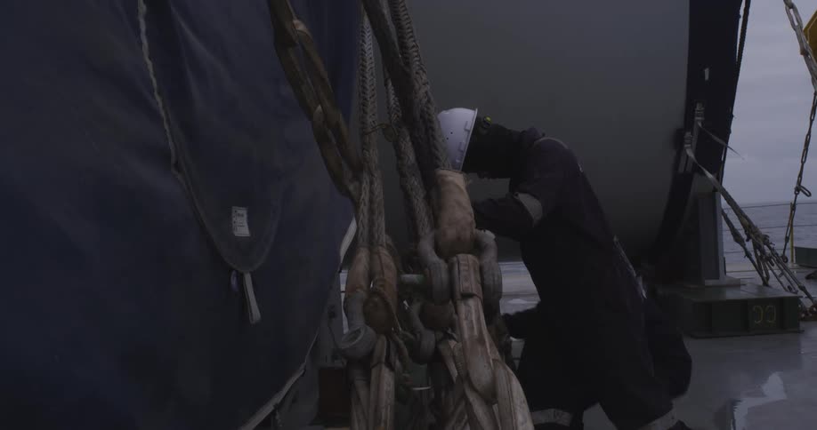 Two maritime workers in hard hats and overalls tighten heavy-duty slings to secure large cargo on the deck of a ship