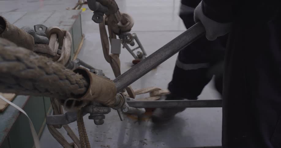 Two industrial workers collaborate to tighten heavy-duty rope rigging with a metal lever on a factory floor