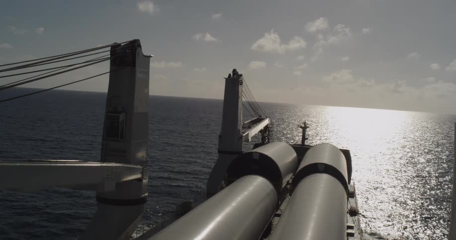 A high angle view shows a large cargo ship transporting wind turbine sections across the vast open ocean towards the sun