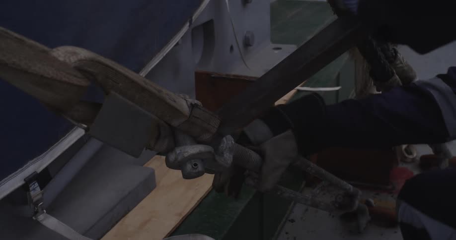 A worker in gloves uses a metal bar to tighten a large industrial turnbuckle and strap