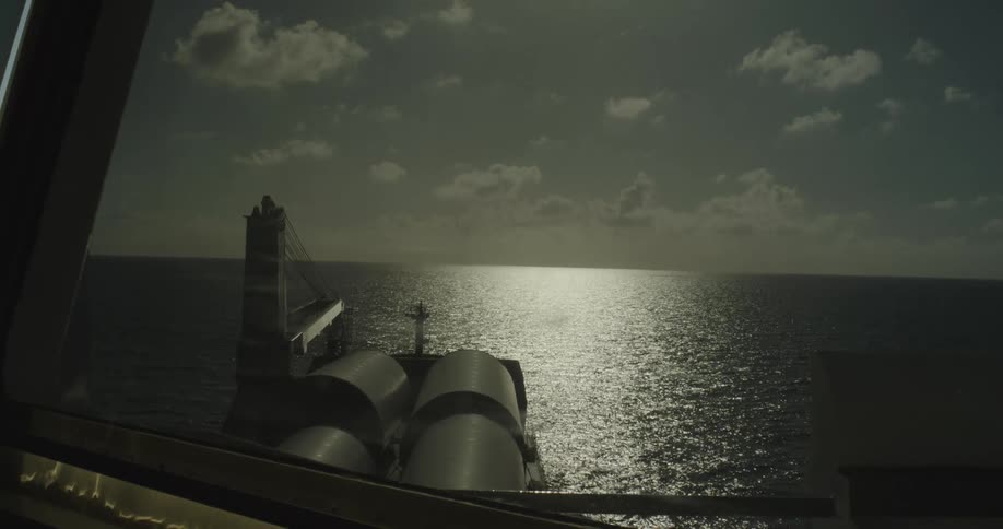 A serene view from a cargo ship's window shows the sun glistening on the vast open ocean as it sails toward the horizon