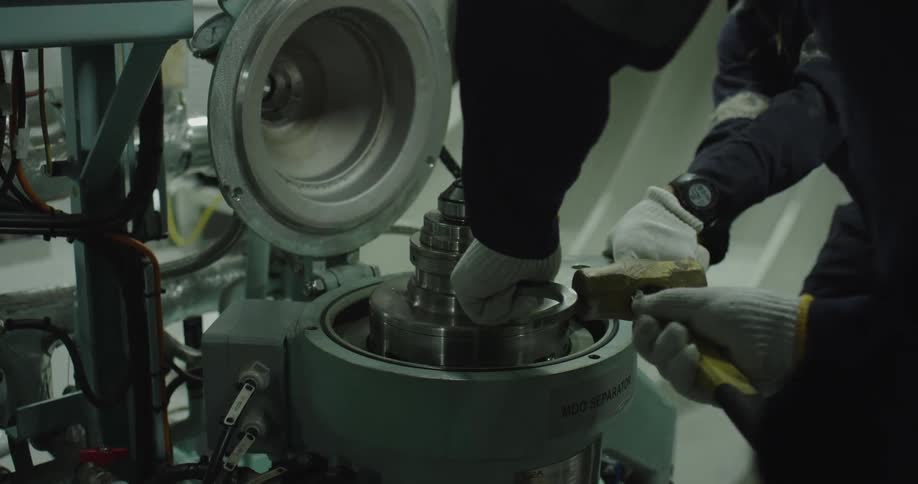 Two marine engineers work together to assemble the internal components of an MDO separator in a ship's engine room, using a wrench and hammer