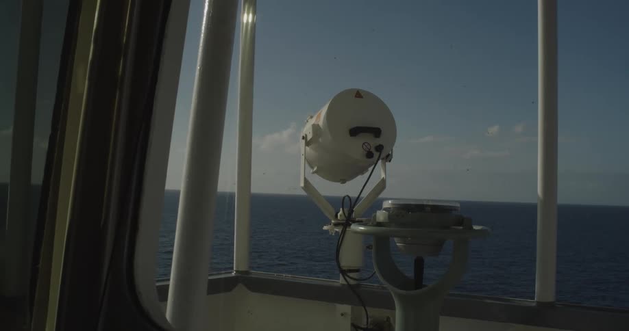A ship's compass and searchlight stand on the bridge overlooking the calm open sea on a sunny day