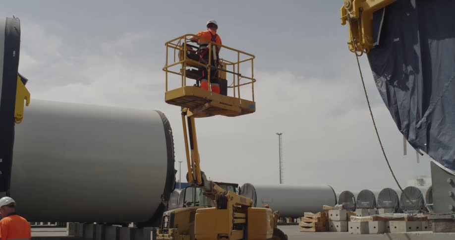 A worker in a safety vest and hard hat operates a yellow cherry picker at an industrial site next to large wind turbine parts