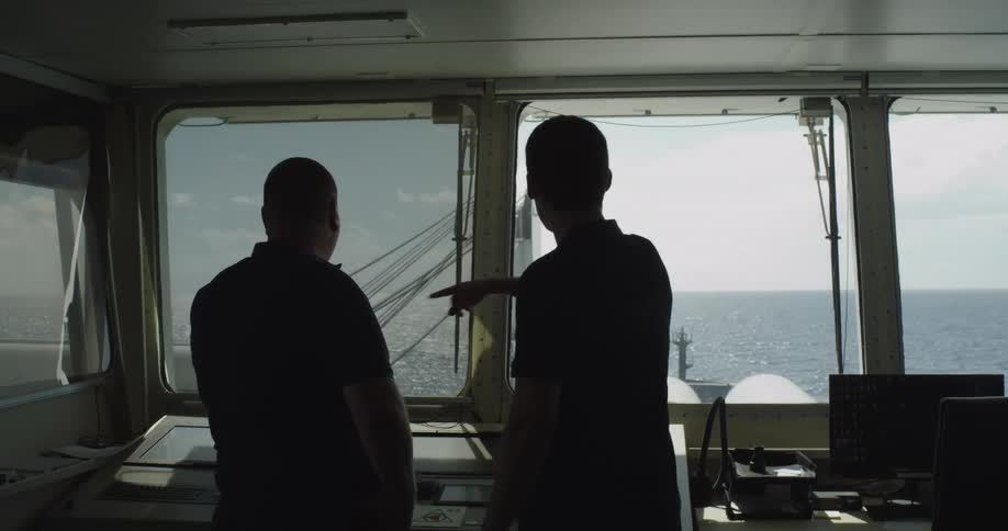 Two crew members are silhouetted on the bridge of a ship as they navigate across the open sea