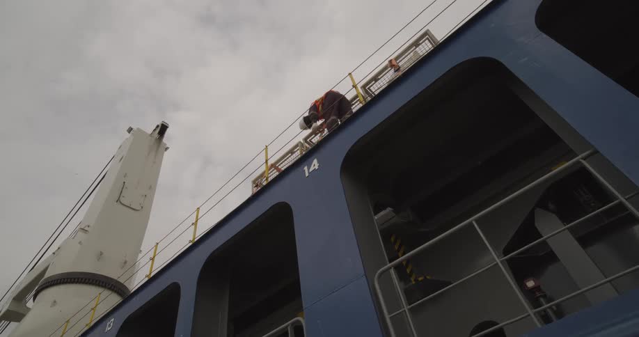 A ship worker performs maintenance high up on the deck of a massive blue cargo vessel under a gray, cloudy sky