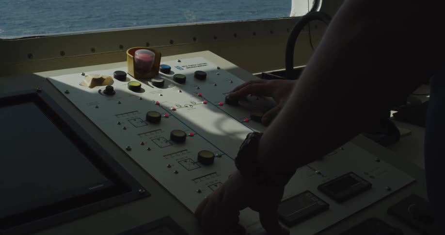 A silhouetted officer's hands operate the control panel on the bridge of a ship sailing on the ocean