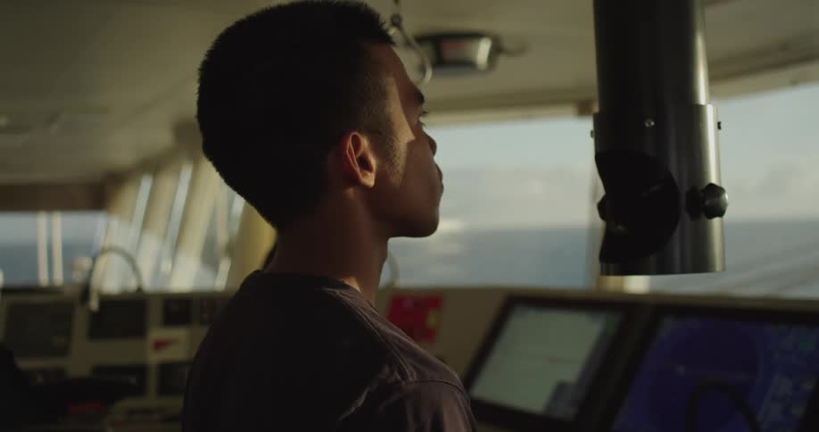A seaman stands on the bridge of a large ship, bathed in golden hour light as he looks out over the open ocean