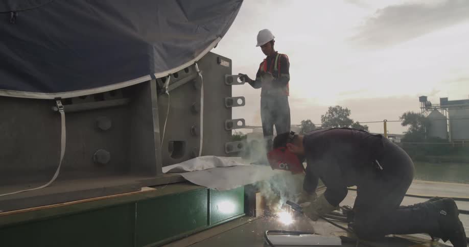 A welder in protective gear works on a large industrial structure while a coworker supervises at an outdoor worksite
