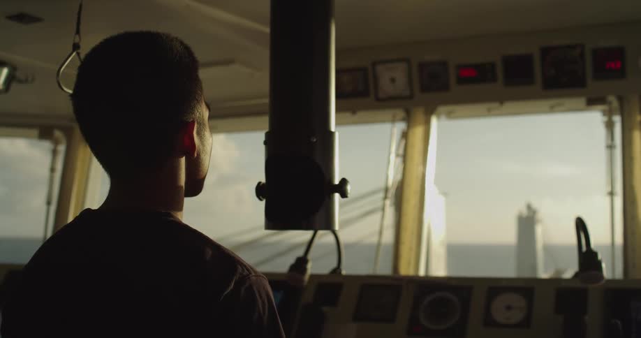 A silhouetted sailor stands on the bridge of a large ship, looking out at another vessel on the ocean during a beautiful sunset