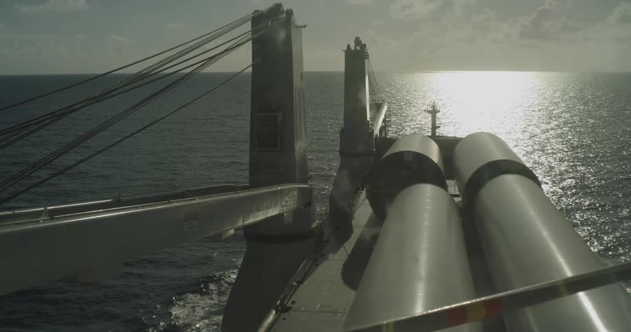 A high-angle view from a cargo ship as it transports large industrial pipes across the open ocean toward the horizon