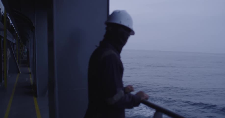 A seaman in a hard hat and balaclava stands by the railing on the deck of a ship, looking out at the calm, gray ocean