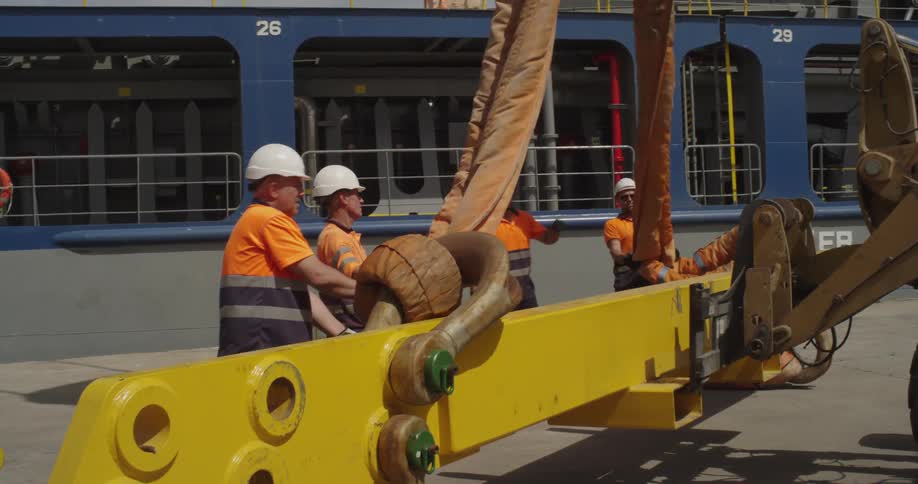 A team of dockworkers in safety gear collaborates to attach heavy lifting straps to a large yellow beam at a port