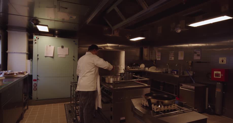 A man in a chef's uniform diligently prepares a meal in a spacious, stainless steel industrial kitchen